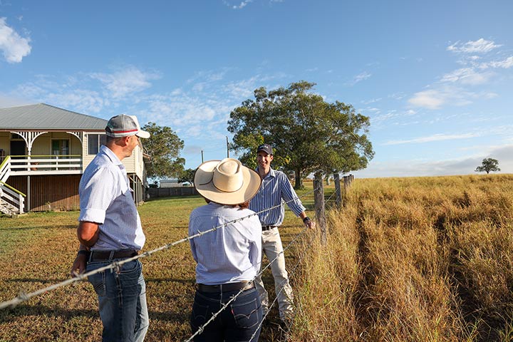 Two people stand next to barb wire fence talking to a man leaning on barb wire fence. The paddock behind them is full of dried off grass and on the left in the background there is a Queenslander home.