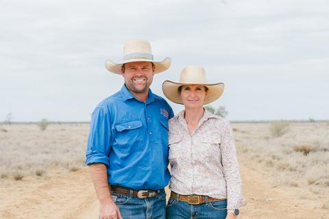 Martin and Beck Eggerling stand on a dusty road, hugging each other smiling at the camera. Martin wears a broad brimmed hat, blue workshirt, brown belt and denim jeans. Beck wears a broad brim hat, light pink work shirt, brown belt and denim jeans. 