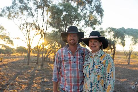 Sam and Julia Burge stand in front of camera smiling as the sun sets behind them amongst the trees